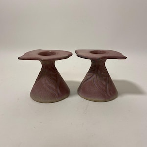 May include: A pair of mauve ceramic candle holders. Each has a square, flared top with a circular candle recess. The base is conical with a textured, leaf-like design. The candle holders are displayed on a white surface.