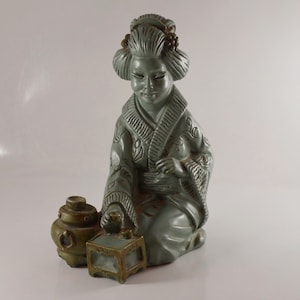 May include: A green ceramic figurine of a woman in a traditional Japanese kimono, holding a small box and sitting next to a green ceramic pot with a lid.