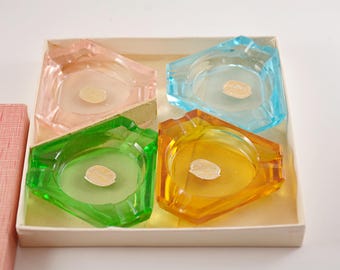 Op de afbeelding: Een set van vier driehoekige glazen asbakken in roze, blauwe, groene en amberkleurige tinten, elk voorzien van een centrale ronde inkeping en een kleine ovale sticker, geplaatst in een vierkante kartonnen doos met een apart roze deksel met textuur.