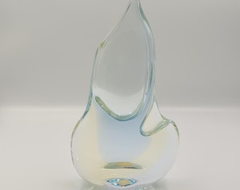 Op de afbeelding: Een heldere glazen vaas met een asymmetrisch, vlamachtig silhouet. De basis heeft zachte, doorschijnende blauwe en lichtgele kleurverlopen die overgaan in het transparante bovenste deel van het sculpturale object.