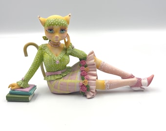 Op de afbeelding: Een gestileerd kattenbeeldje met een gele huid, met een groene hoofddoek met stippen, een groen shirt met lange mouwen en ruches, en een roze geruite rok versierd met kleine roze en gele bloemen. De kat rust met één hand op drie boeken.