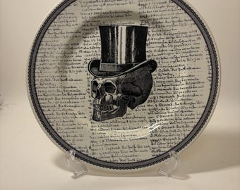 May include: A decorative plate featuring a black and white skull wearing a top hat. The plate has a border with a delicate pattern and a background of handwritten script. A unique gothic home decor item.