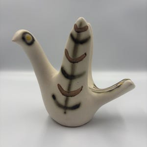 May include: A ceramic bird figurine in an off-white matte finish. The bird features stylized brown and black painted markings on its upright wing and tail, with a simple circular eye detail containing a yellow center on its head.