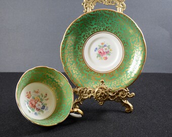 May include: A green and gold teacup and saucer with a floral design. The saucer is on a decorative stand.