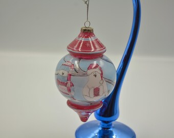 Op de afbeelding: Een ronde, uivormige kerstbal van glas met een lichtblauwe achtergrond, voorzien van illustraties van een sneeuwpop en een ijsbeer met een rode muts. De hanger heeft een rode boven- en onderkant met witte accenten en een zilverkleurig koord.