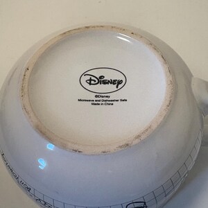 Puede incluir: Taza Disney de cer&aacute;mica blanca con un patr&oacute;n de cuadr&iacute;cula negra y bocetos de dibujos animados. La base de la taza presenta el logotipo de Disney y texto que indica que es apta para microondas y lavavajillas, fabricada en China.