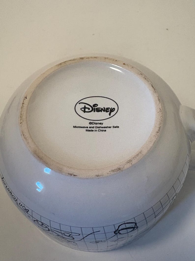 Puede incluir: Taza Disney de cer&aacute;mica blanca con un patr&oacute;n de cuadr&iacute;cula negra y bocetos de dibujos animados. La base de la taza presenta el logotipo de Disney y texto que indica que es apta para microondas y lavavajillas, fabricada en China.