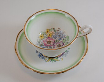 May include: A white teacup and saucer set with a floral design. The cup and saucer have a green border and gold trim. The inside of the cup features a bouquet of pink, yellow, and blue flowers. The saucer has a matching floral design.