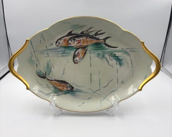 Op de afbeelding: Een ovale keramische serveerschaal met een geschulpte rand en goudkleurige handvatten. Het witte oppervlak is voorzien van een handgeschilderde illustratie van vier vissen die in blauw en groen water zwemmen, met abstracte lijndetails.