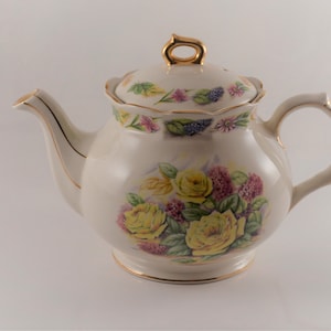 May include: A white ceramic teapot with a floral design featuring yellow roses, pink and purple flowers, and green leaves. The teapot has a gold trim around the lid and base.