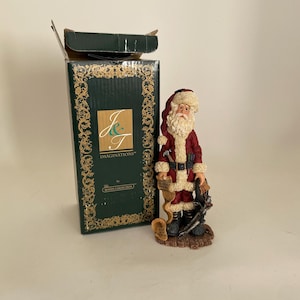 May include: A red and white Santa Claus figurine holding a scroll and tools. The figurine is standing on a wooden base. The box is green with gold trim and the words "J & T Imaginations" and "The Royal Collection".
