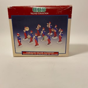 May include: A Lemax Village Collection box featuring a porcelain village accessory. The box has a red top and a blue background with a band of musicians in blue uniforms and red hats. The text "PORCELAIN VILLAGE ACCESSORY" is printed on the box.