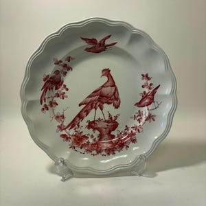 May include: White ceramic plate with a scalloped edge, featuring a red bird and floral design. The plate has a detailed illustration of a pheasant and other birds, with a decorative border. The plate is displayed on a clear stand.