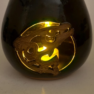 May include: A dark green, glossy ceramic candle holder with a carved wooden fish design. The fish is set within a circular cutout, and a warm yellow light glows from inside, creating a cozy ambiance. The candle holder has a rounded shape.