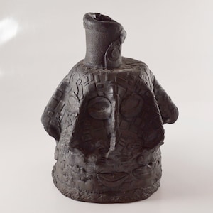 May include: A black ceramic vase with a face-like design. The vase has a wide base and a narrow neck. The face is made up of geometric shapes and patterns.