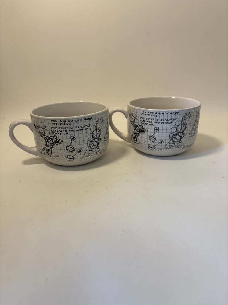 Puede incluir: Dos tazas de cer&aacute;mica blanca con asas. Cada taza presenta una ilustraci&oacute;n de dibujos animados en blanco y negro y un texto manuscrito que dice "TRY FOR DAINTY HAND POSITIONS. THE SHIRT IS SLIGHTLY STARCHED AND USUALLY STICKS UP."