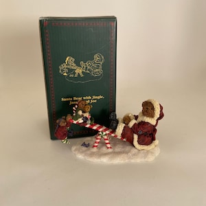 May include: A Christmas figurine of a Santa Bear on a seesaw with other bear figures. The bears wear festive outfits, with a red and white striped seesaw. The figurine is on a snowy base, with a green box in the background.