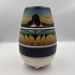 May include: A ceramic vase featuring a painted desert landscape with a dark silhouette of a mesa against a blue and orange gradient sky. The middle section displays a geometric bridge pattern in teal and yellow, while the base is solid black and cream.