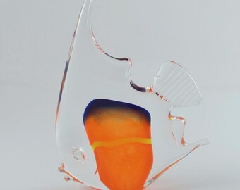 Op de afbeelding: Een sculptuur van helder glas in de vorm van een maanvis met een feloranje lichaam, een donkerblauw bovengedeelte en een dunne horizontale gele streep. Het glazen figuur heeft een hoge rugvin, een klein oog en een gestructureerde staartvin.