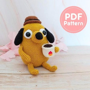 Könnte beinhalten: Eine gehäkelte gelbe Hundefigur mit schwarzen Ohren und einem braunen Hut, die eine kleine Kaffeetasse mit einem Flammendesign hält. Die Figur hat große, runde Augen. Das Bild enthält einen rosa Kreis mit dem Text "PDF Pattern".