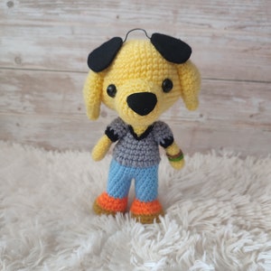 May include: A crocheted yellow dog toy with black ears, a black nose, and black eyes. The dog is wearing a gray shirt and blue jeans with orange accents.