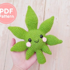 Puede incluir: Un juguete de peluche en forma de hoja verde tejido a ganchillo con una cara sonriente, ojos de botón negros y mejillas rosas. El juguete se sostiene en una mano. El texto "PDF Pattern" está en un círculo rosa en la esquina superior izquierda.