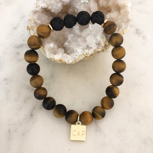May include: A beaded bracelet featuring tiger's eye and black lava stone beads. The bracelet includes a gold-colored charm with the letters "C&P". The bracelet is displayed on a white surface.