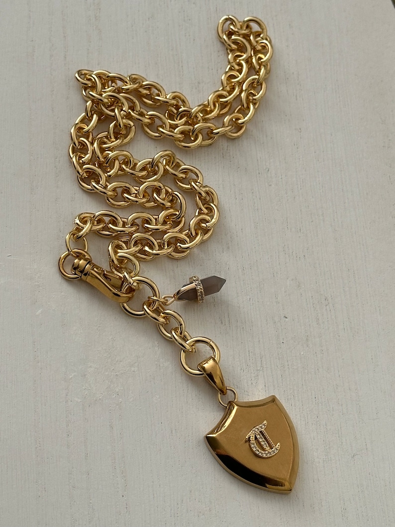 Puede incluir: Collar de cadena dorada con un colgante en forma de escudo. El colgante presenta la letra "C" adornada con peque&ntilde;os cristales. Tambi&eacute;n se ve un peque&ntilde;o cristal puntiagudo y un cierre de mosquet&oacute;n.