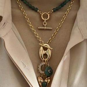 Op de afbeelding: Een gelaagde kettingenset met een bovenste streng donkergroene kralen met een goudkleurige knevelsluiting en een onderste goudkleurige ketting met een grote schakel, een halve maan bedel, een groene steen en een getextureerde gouden hartbedel.