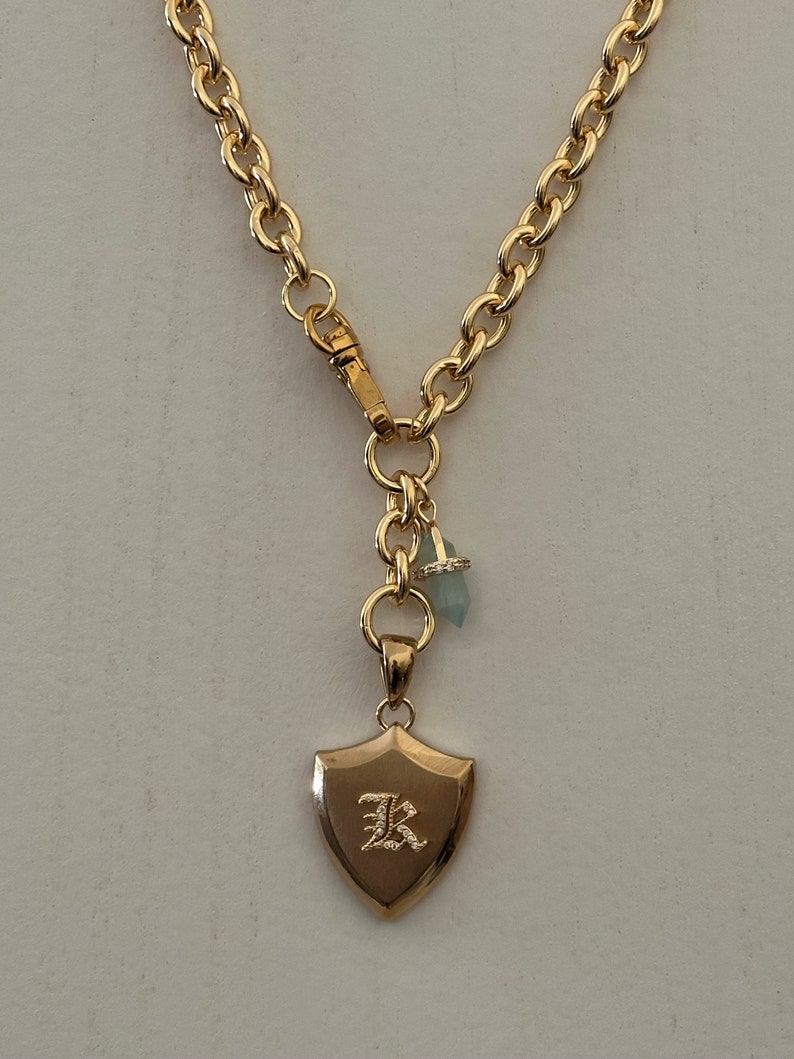 Puede incluir: Collar dorado con un colgante en forma de escudo con la letra "K". La cadena tiene eslabones ovalados grandes y un cierre de mosquet&oacute;n. Un peque&ntilde;o dije con una piedra azul claro y un anillo con diamantes cuelgan de la cadena.