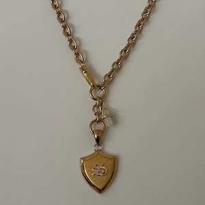 Puede incluir: Collar dorado con un colgante en forma de escudo. El colgante presenta un emblema detallado. La cadena es de estilo eslab&oacute;n cl&aacute;sico. Una peque&ntilde;a piedra transparente est&aacute; unida a la cadena cerca del cierre. El collar se muestra sobre un fondo blanco.