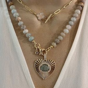 May include: A gold chain necklace with a heart-shaped pendant featuring a green gemstone surrounded by smaller diamonds. The necklace is layered over a beaded necklace with light green and brown beads.
