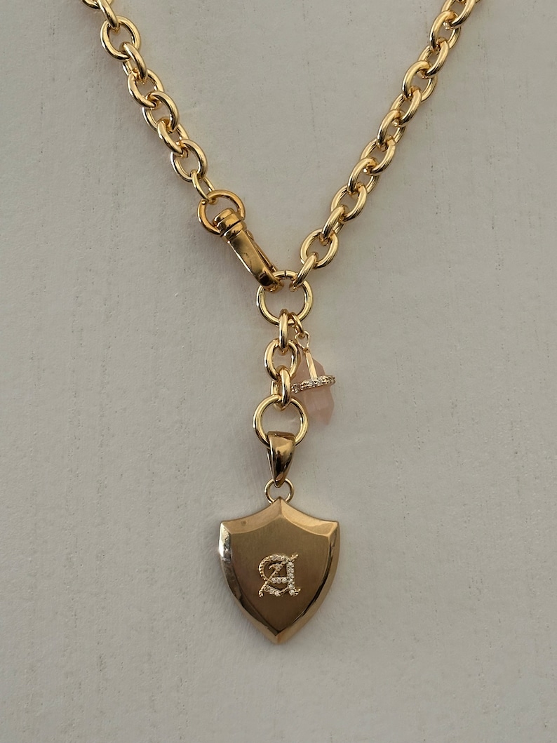 Puede incluir: Collar dorado con un colgante en forma de escudo. El colgante presenta la letra "A" en una fuente decorativa y est&aacute; adornado con peque&ntilde;os cristales. El collar tiene una cadena gruesa y un cierre de mosquet&oacute;n.