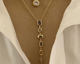 Pode incluir: Dois colares de ouro. O colar superior tem uma corrente com um pingente redondo com uma pedra transparente. O segundo colar tem uma corrente com uma série de pedras preciosas coloridas em várias formas, incluindo um retângulo, um oval e uma lágrima.