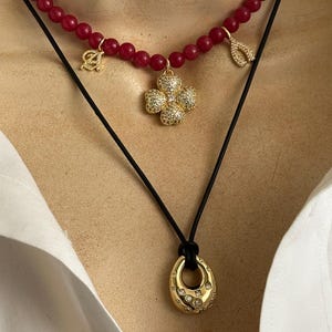 May include: A layered necklace set featuring a strand of round red beads with a gold-tone letter A charm, a gold-tone wishbone charm, and a central gold-tone four-leaf clover pendant with clear stones, paired with a black cord and gold-tone teardrop pendant.