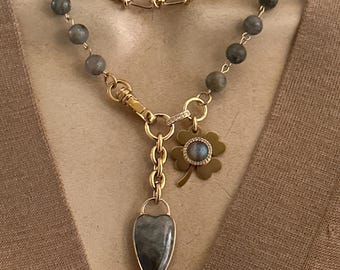May include: A layered gold-toned necklace with a heart-shaped pendant and a four-leaf clover charm. The necklace features gray gemstone beads and a chain link design. The pendant and clover charm have a gemstone center.