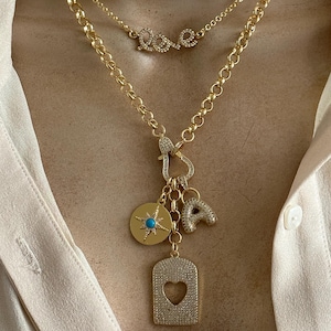 May include: A gold chain necklace with a charm pendant featuring a compass, a heart-shaped tag, and a letter 'A'. The necklace also includes a smaller chain with a 'love' charm.