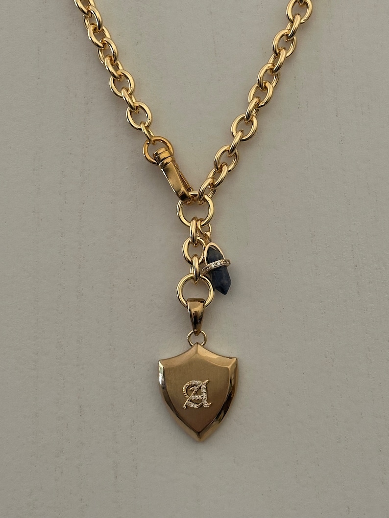 Puede incluir: Collar dorado con un colgante en forma de escudo con la letra "A". El collar tiene una cadena gruesa y un cierre de broche. Tambi&eacute;n hay un peque&ntilde;o dije de piedra preciosa azul adjunto.