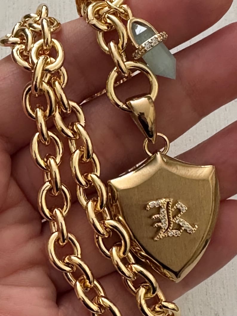Puede incluir: Collar dorado con un dise&ntilde;o de cadena gruesa. El collar presenta un colgante de escudo con la letra "R" adornada con peque&ntilde;os cristales. Tambi&eacute;n se adjunta un colgante de cristal verde claro con un anillo con incrustaciones de diamantes.
