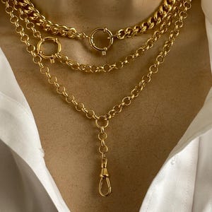 May include: Three gold necklaces of varying styles are displayed. The top necklace is a thick curb chain, the middle is a delicate chain with a large ring clasp, and the bottom has a long drop with a lobster clasp. The necklaces are worn over a white shirt.
