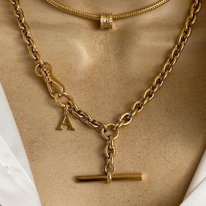 May include: A gold chain necklace with a t-bar clasp and a letter 'A' charm. The necklace is layered over a thinner gold chain with a round charm.