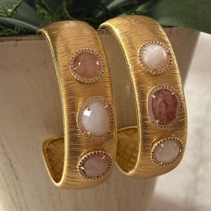 May include: Two gold-toned, textured cuff bracelets. Each bracelet features three oval-shaped, light-colored gemstones, each surrounded by a ring of small, clear stones. The bracelets are displayed against a neutral background.