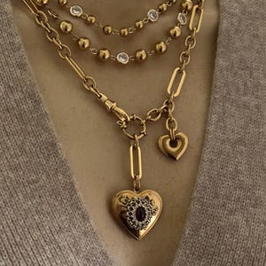 May include: A layered gold necklace set. The top layer features small gold beads with clear accents. The middle layer has a chain link design. The bottom layer has a heart-shaped locket with a dark center stone and a small heart charm.