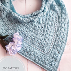 Peut inclure: Un châle triangulaire en crochet bleu clair avec un motif ajouré. Le châle est plié et drapé, mettant en valeur sa texture et son motif. Les mots "PDF PATTERN" et "US TERMS" sont visibles en bas à gauche.