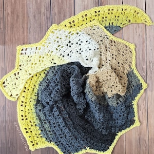 Può includere: Uno scialle all'uncinetto fatto a mano nei toni del giallo, bianco, beige e grigio. Lo scialle ha un bordo smerlato e un design a spirale. Lo sfondo è una superficie di legno.