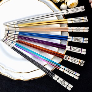 Könnte beinhalten: Ein Set mit zehn Paar Essstäbchen mit unterschiedlichfarbigen Griffen und personalisierten Gravuren. Die Essstäbchen sind fächerförmig auf einem weißen Teller mit goldfarbenem Rand angeordnet. Die Gravuren beinhalten Namen, Daten und Initialen.