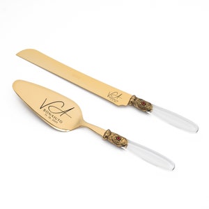 May include: A gold-toned cake knife and server set featuring clear acrylic handles with ornate, antique-style brass-colored metal accents and a small red stone inlay. The blades are engraved with the text VA BONAIUTO and the date 11. 10. 2023.