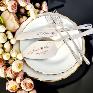 May include: A wedding cake serving set with a knife and server, along with two forks, resting on a white plate with a gold rim. The knife and server are engraved with names and a date. Pink and white flowers are in the background.