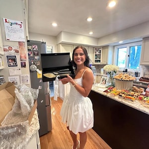 A smiling woman in a white bridal dress holding an open premium black magnetic gift box with a personalized champagne gold wedding cake cutting set. Shown in a kitchen during a celebratory bridal shower with a large food spread.