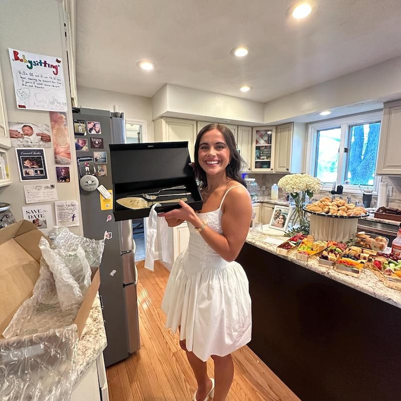 A smiling woman in a white bridal dress holding an open premium black magnetic gift box with a personalized champagne gold wedding cake cutting set. Shown in a kitchen during a celebratory bridal shower with a large food spread.